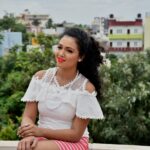 Home A cheerful woman sitting outdoors in a white lace top against a cityscape backdrop.