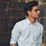 Home A stylish young man in glasses leans against a brick wall wearing a casual light blue shirt.